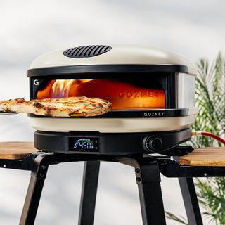 Gozney arc pizza oven on a stand in front. of a white wall, with pizza being placed into the lit oven