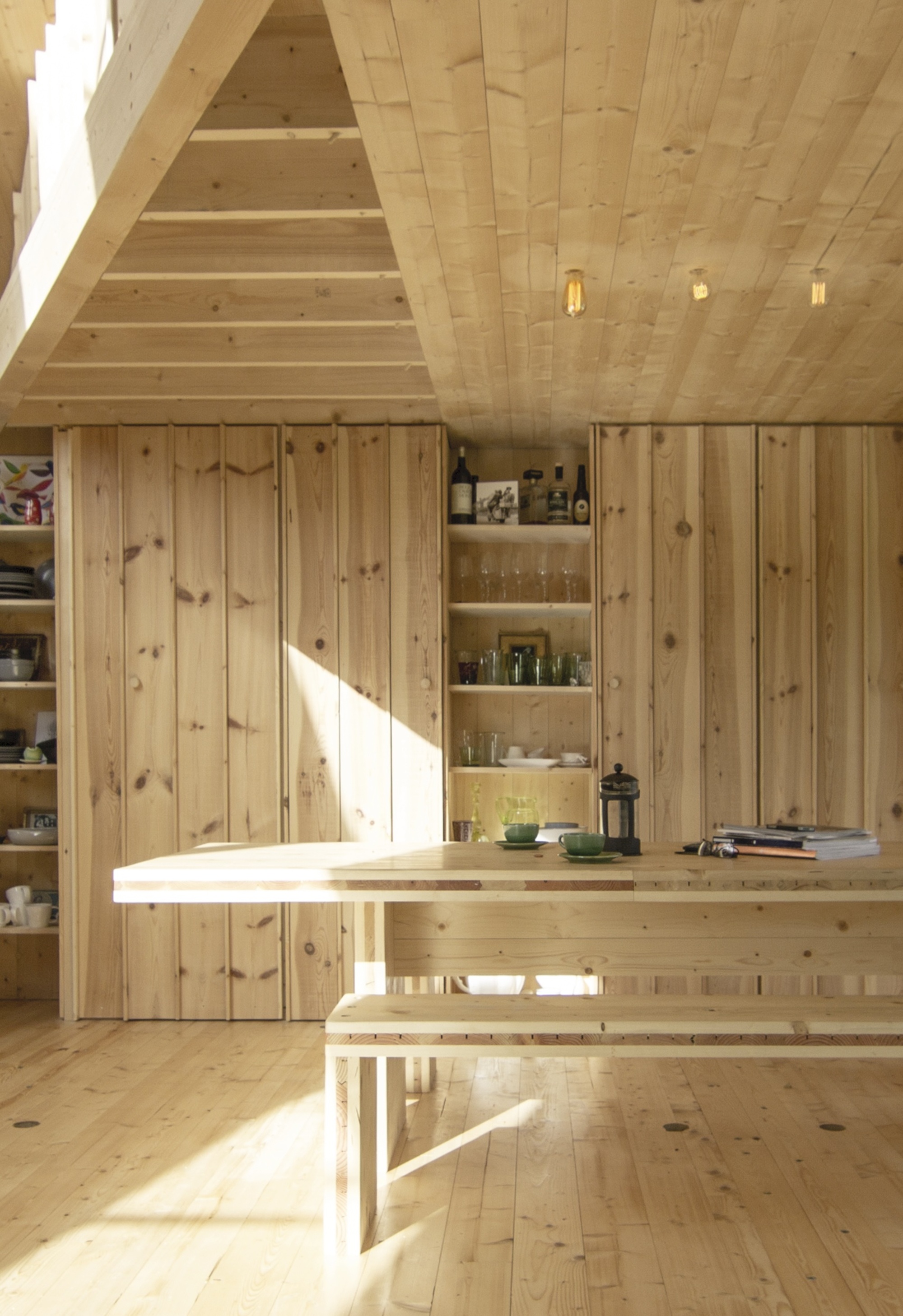 scandinavian timber clad dining room with timber benchseat, shelving styled with glassware, plates, wine, and cafeteria and cups on the table