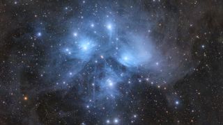 The stars of the Pleiades open cluster shine brightly against a more distant starfield, while surrounded by blue nebulosity.