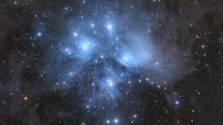 The stars of the Pleiades open cluster shine brightly against a more distant starfield, while surrounded by blue nebulosity.