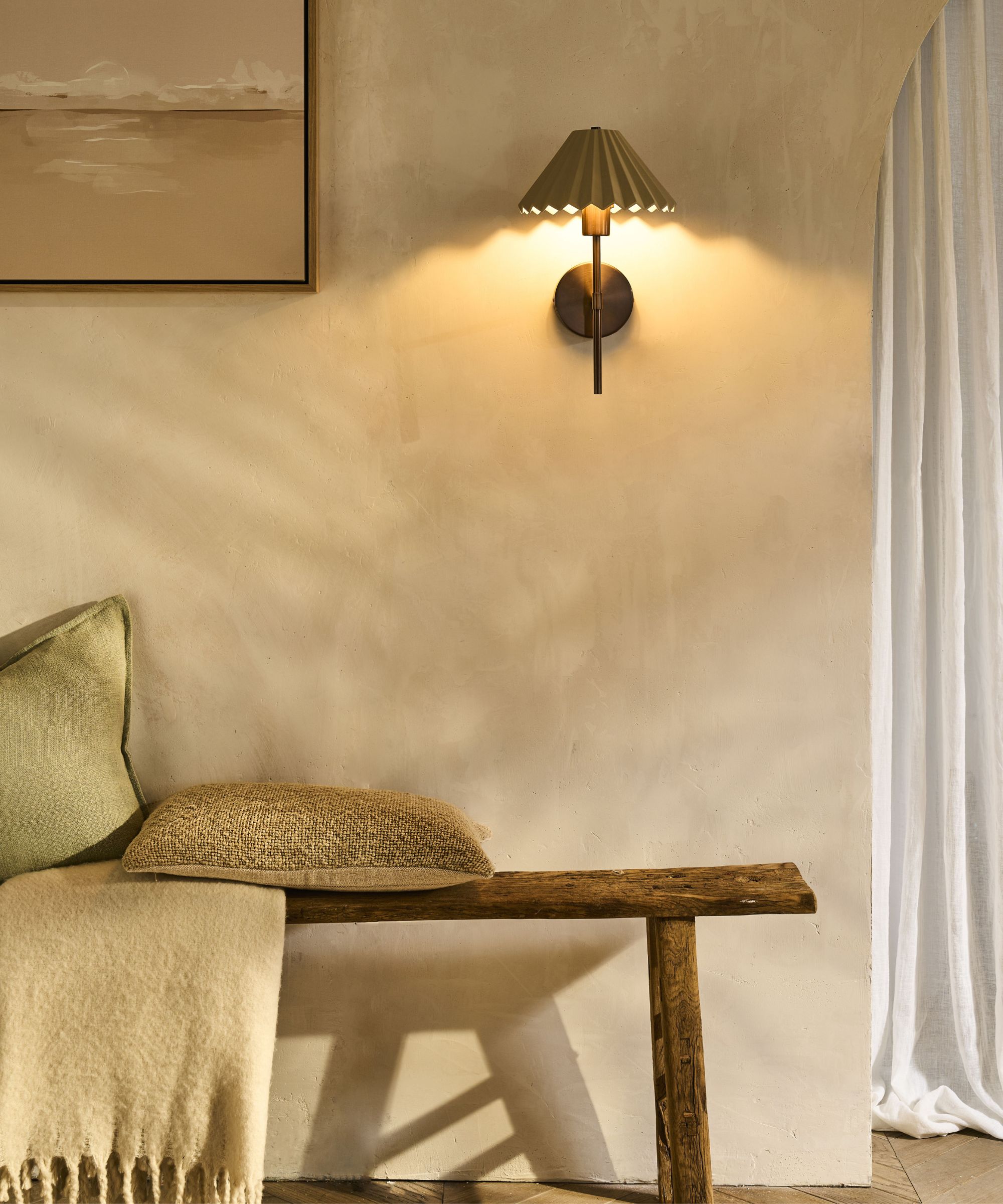 A rustic, softly lit corner featuring a wooden bench with textured cushions and a wool throw. The warm glow of a pleated wall sconce enhances the plastered wall&amp;rsquo;s natural, tactile finish