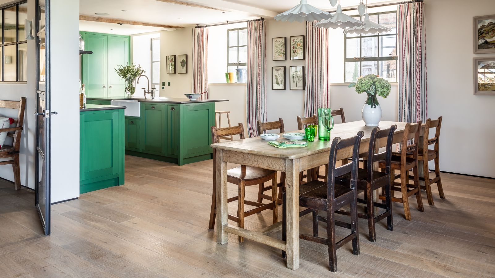A country kitchen diner with green kitchen cabinets in the background, and a six-chair wooden dining table in the foreground. 