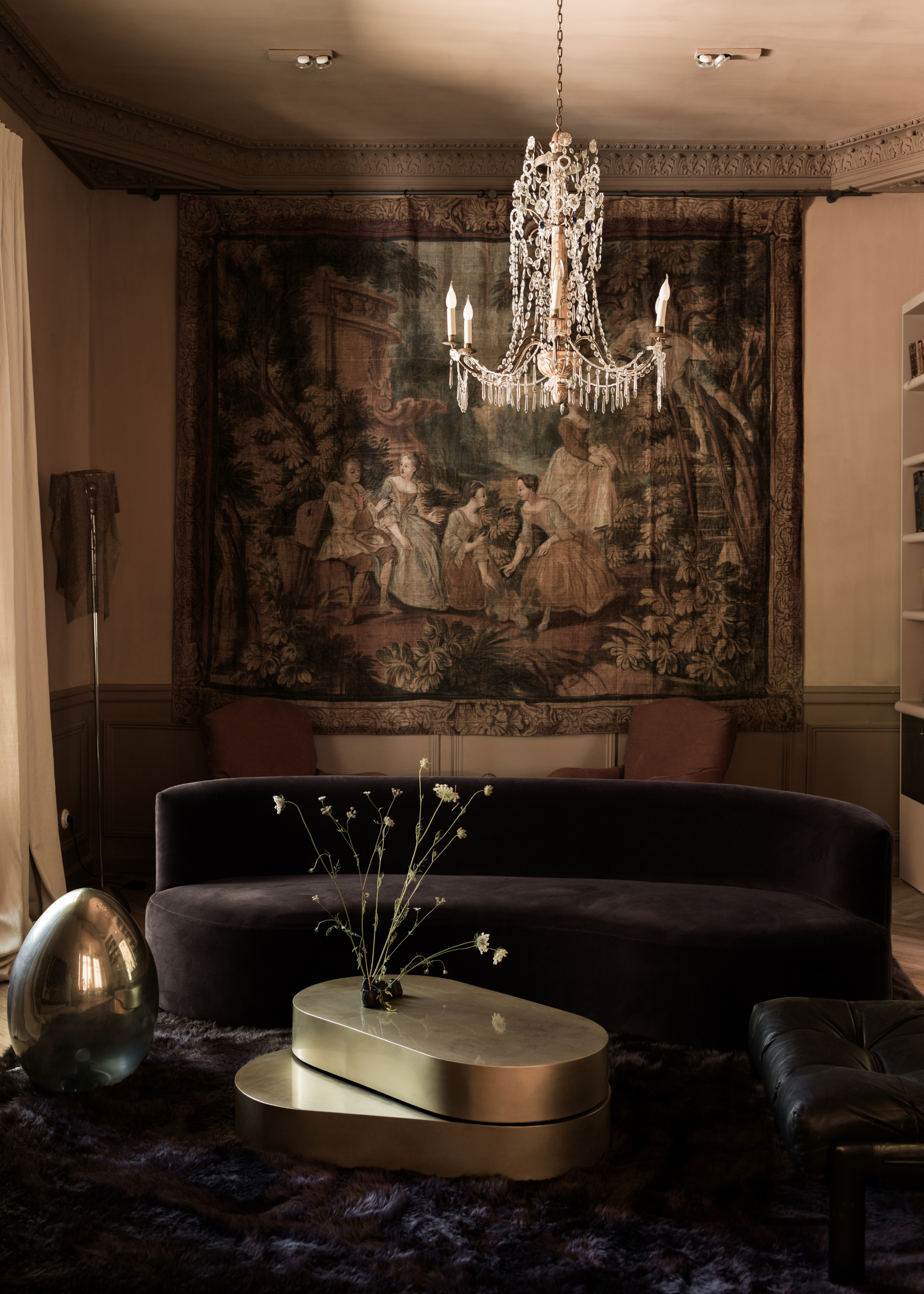 A living room with a chandelier, a hanging mural, a black sofa, a gold coffee table, a silver egg sculpture, and a chainmail lamp