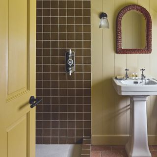 bathroom with brown square tiles in shower and cream painted wood panel walls with pedestal sink and terracotta brick flooring