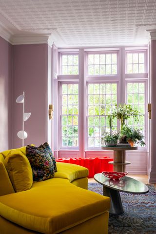 A living room painted in a pale purple with large windows and a yellow sofa