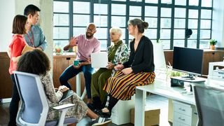 A diverse group of people chatting in a modern office. One woman is an amputee with a prosthetic leg, sat on a desk.