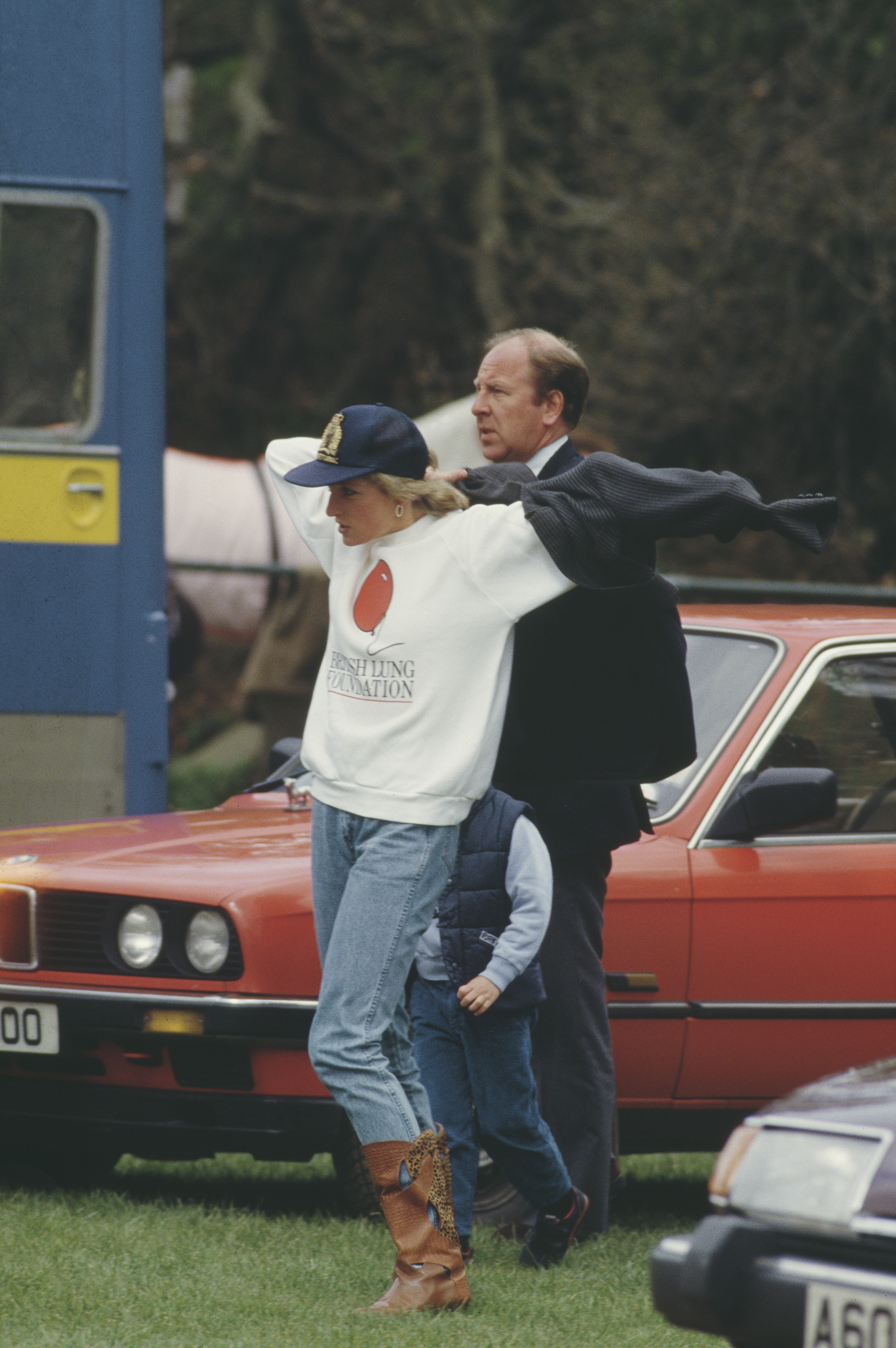 Princess Diana putting a blazer on standing in front of a red car and wearing jeans and a white sweatshirt