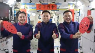 three astronauts in blue sweat suits hold up red paper cuttings in a white rectangular space station cabin