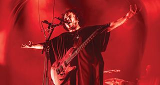 Manuel Gagneux of Zeal & Ardor pictured onstage, arms outstretched with his Aristides electric guitar, on a stage light in red