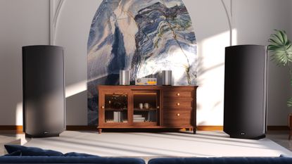 A pair of Quad ESL 2912X speakers in a brightly lit room on either side of a wooden sideboard with Quad audio components on top of it