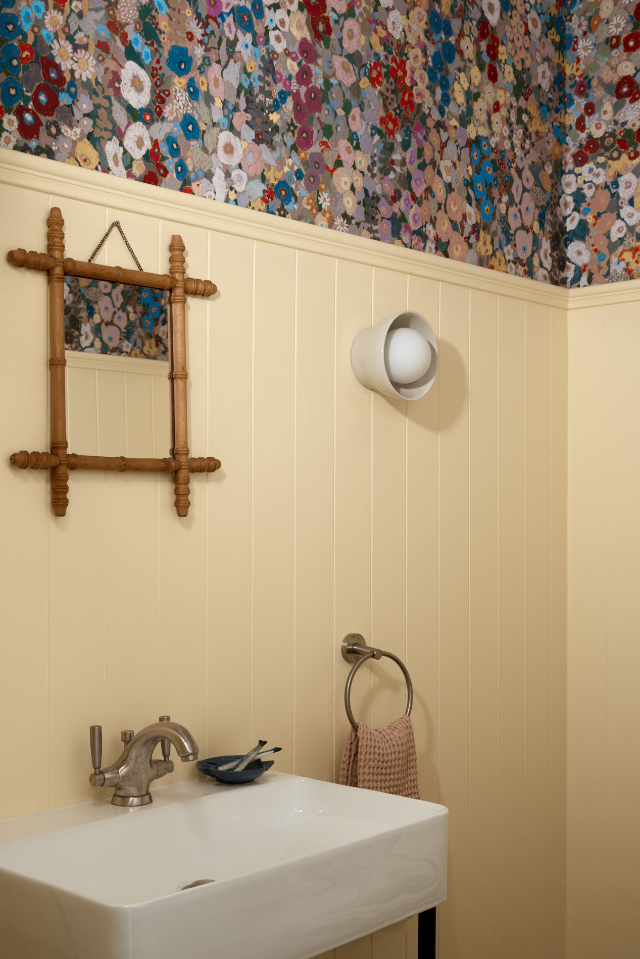 small bathroom with butter yellow wall panelling, a wall sconce, bamboo mirror, white sink with hand towel hanging beside, and hollyhock wallpaper above a dado rail