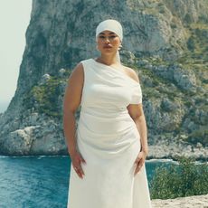  model wears white dress and white headband while standing in front of a gorgeous ocean and rock mass 