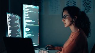 Female developer carrying out software security flaw remediation tasks on desktop computer in dimly-lit room with two monitors.