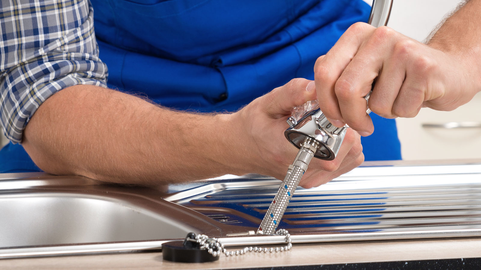 Close up of man in blue overalls fitting a Chrome mixer tap to a stainless steel kitchen sink