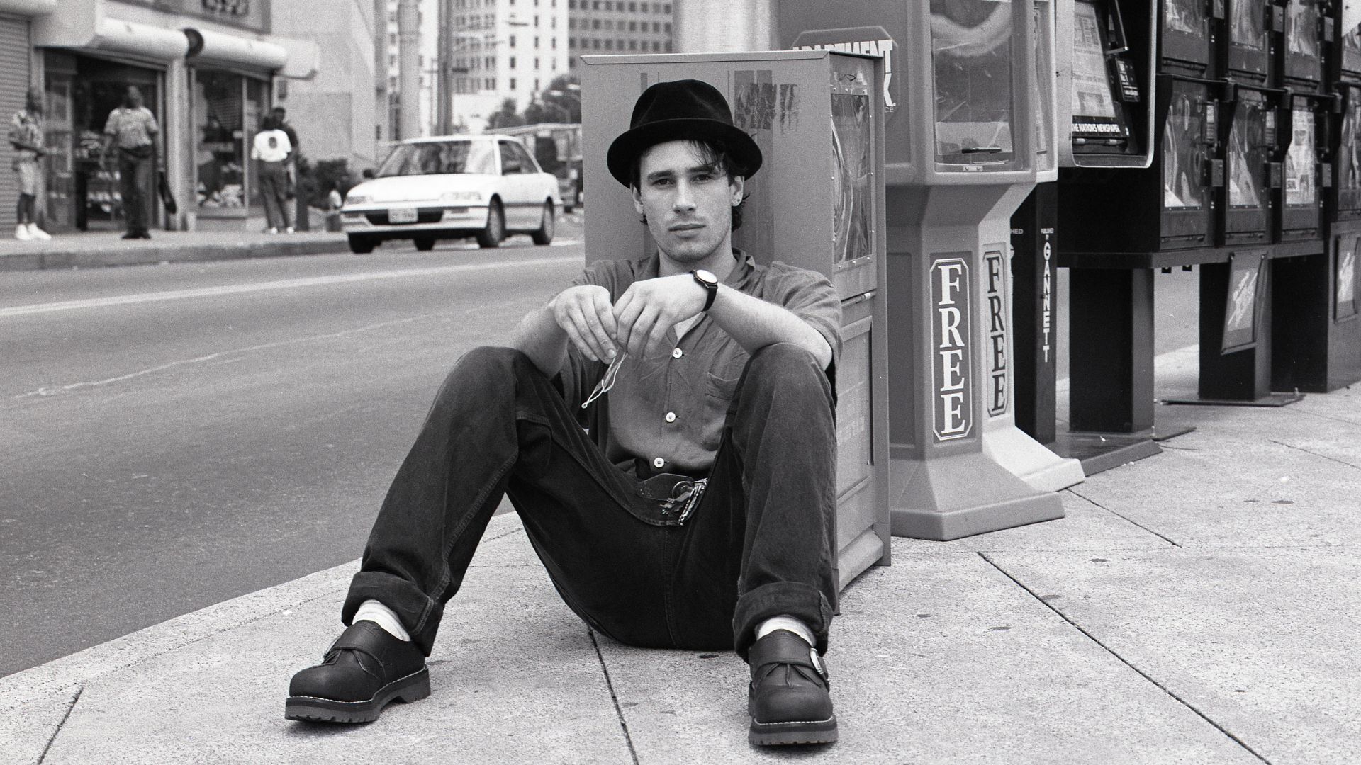 a black and white image of Jeff Buckley who has been photographed posing whilst sitting on the pavement next to a busy street in America. He is leaning against a newspaper stand