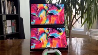 Dual monitors displaying vibrant abstract art with colorful swirls, placed vertically on a wooden table. A gaming controller is between them, with books and a plant in the background.