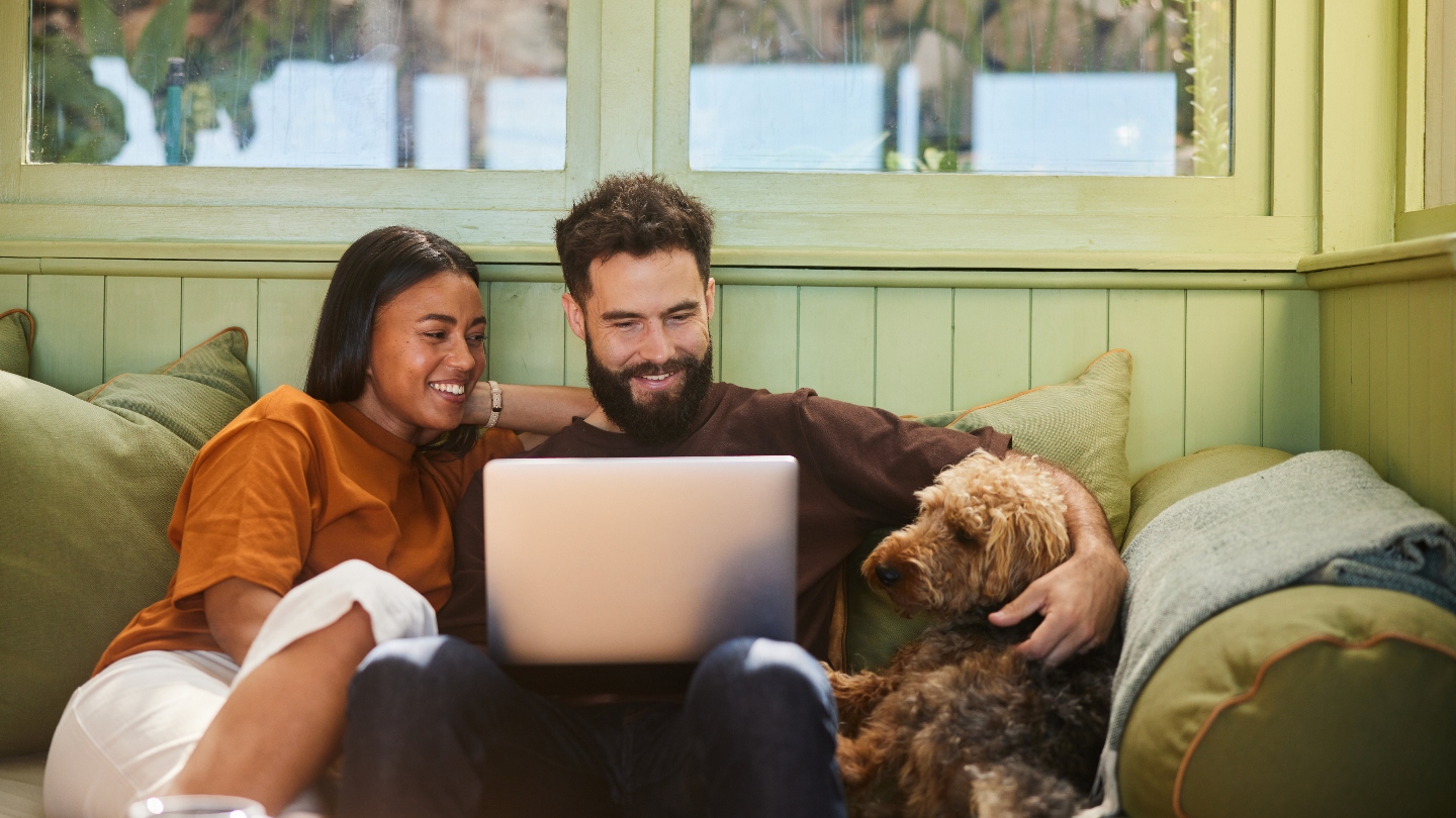 Couple sitting on a couch smiling at a laptop on the man&#039;s lap with a dog next to them