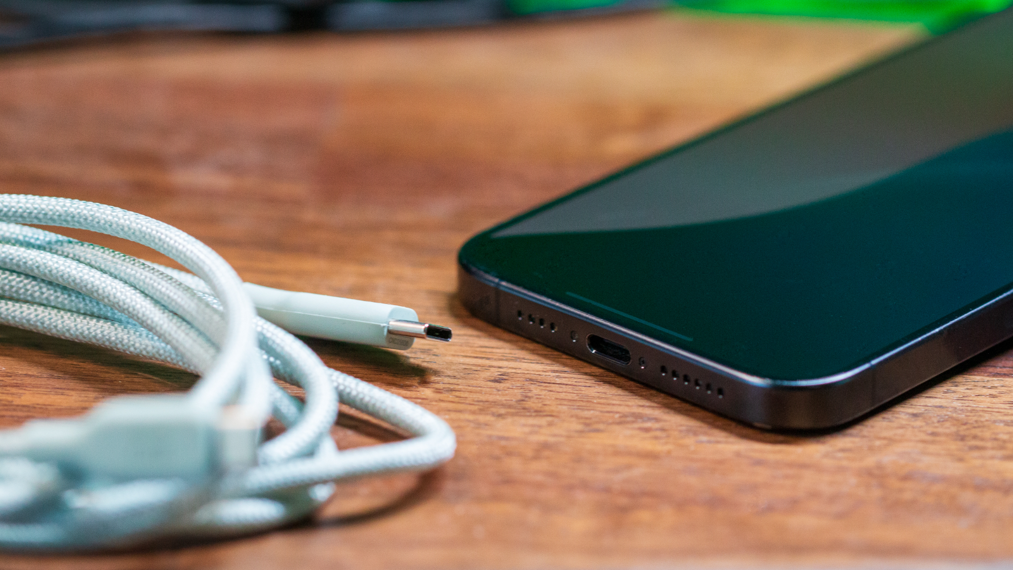 A USB-C cable and iPhone 15 Pro Max are placed on a wooden table, showing the iPhone's USB Type-C port.