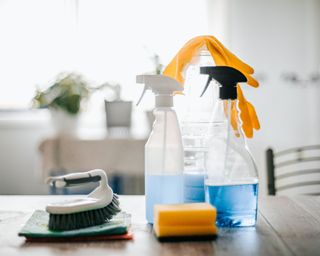 Set on cleaning supplies on a kitchen table including two spray bottles, yellow gloves and sponge