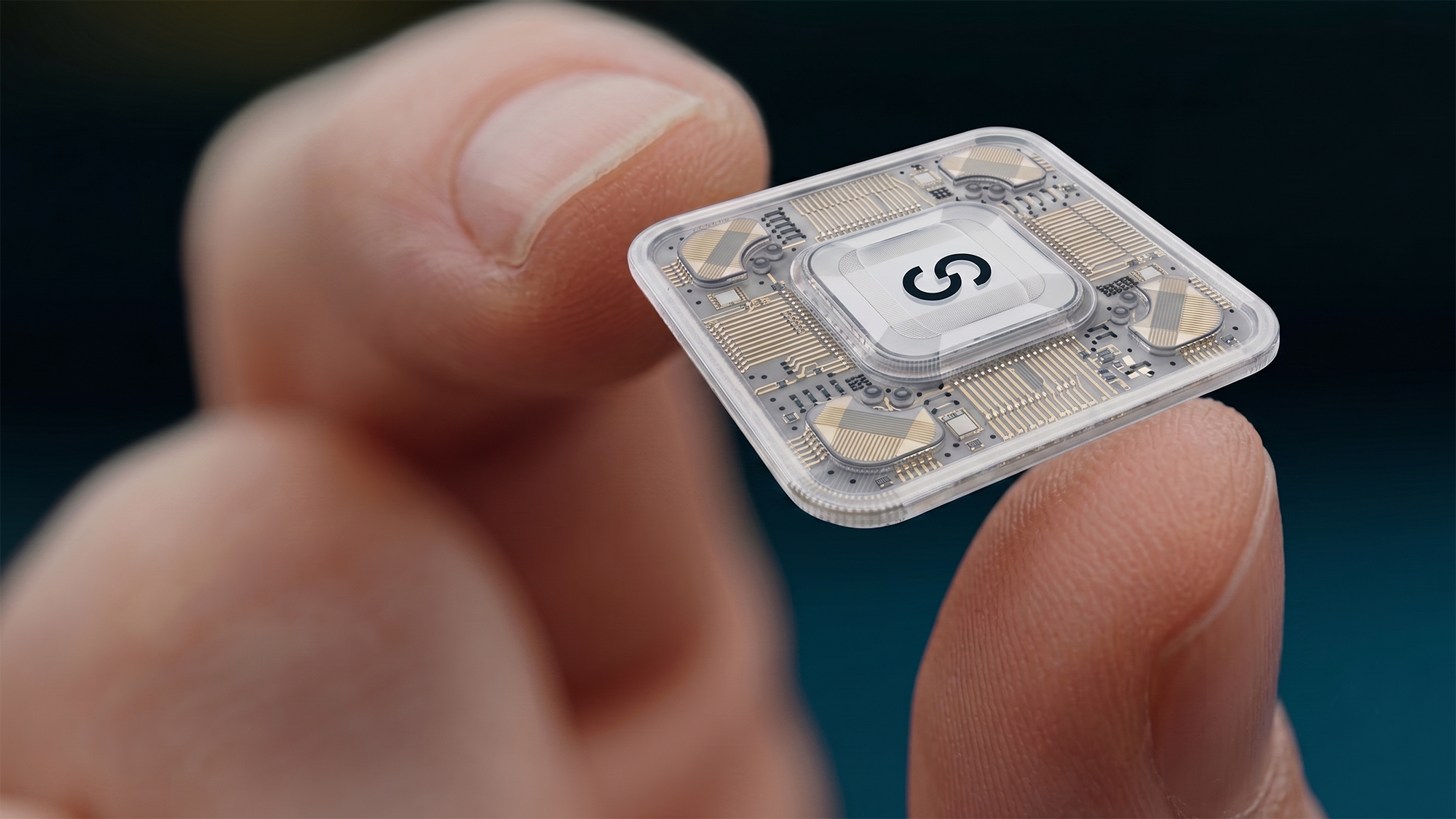 A close up of someone's hand holding a small electronic circuit chip with the letter S on it in the middle