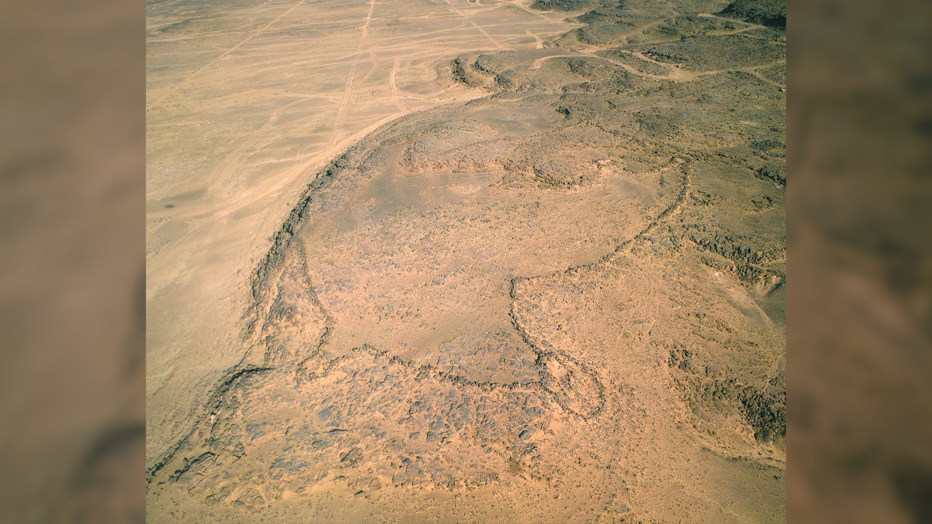 An aerial view of a desert kite. We see a large stone outline with a roundish shape in the desert.