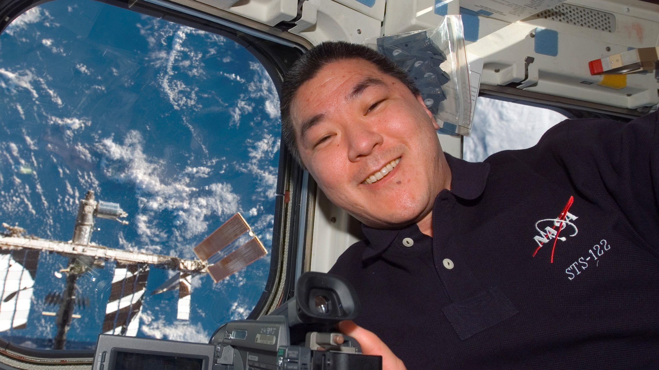 Former NASA space shuttle astronaut Dan Tani aboard a NASA Space Shuttle mission to the International Space Station. The ISS can be seen outside a window while Tani smiles in front of it.
