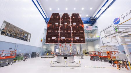 inside a white hangar with a large ceiling sits a metal cylinder from which extend three long rectangular solar panels, all pointing towards the ceiling