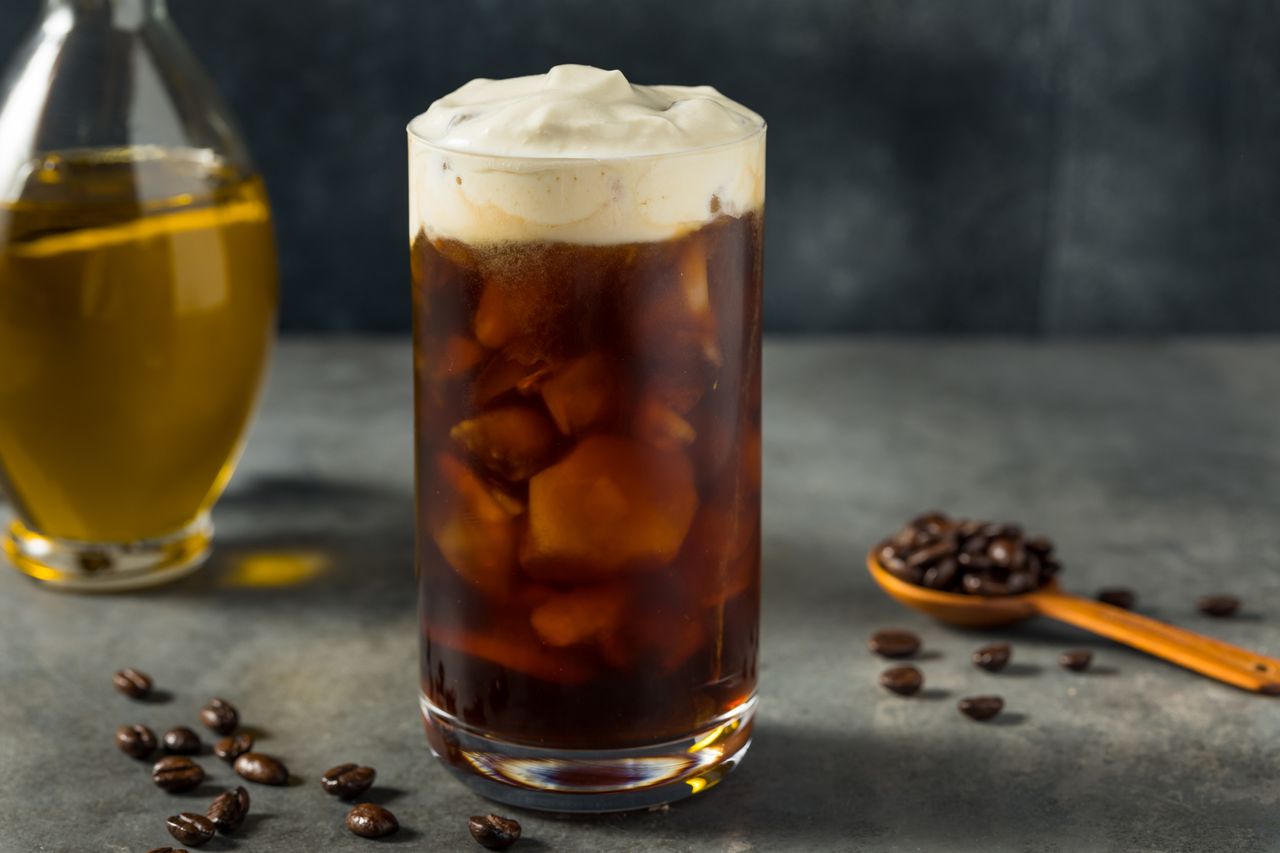 A Oleato coffee - an olive oil coffee - on a countertop w coffee beans and a bottle of olive oil