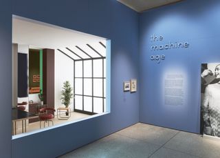 A blue interior for the exhibition, with writing on the wall "The machine age". Through an opening, we see a dining room set up, with a black desk and a reading chair, with a wall to ceiling windows on the other side.