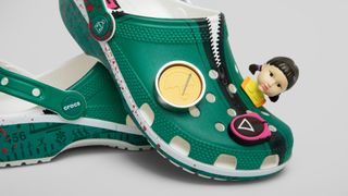 Squid Game Crocs piled on top of each other against a gray background, with Red Light Green Light Girl Jibbitz staring out into the foreground
