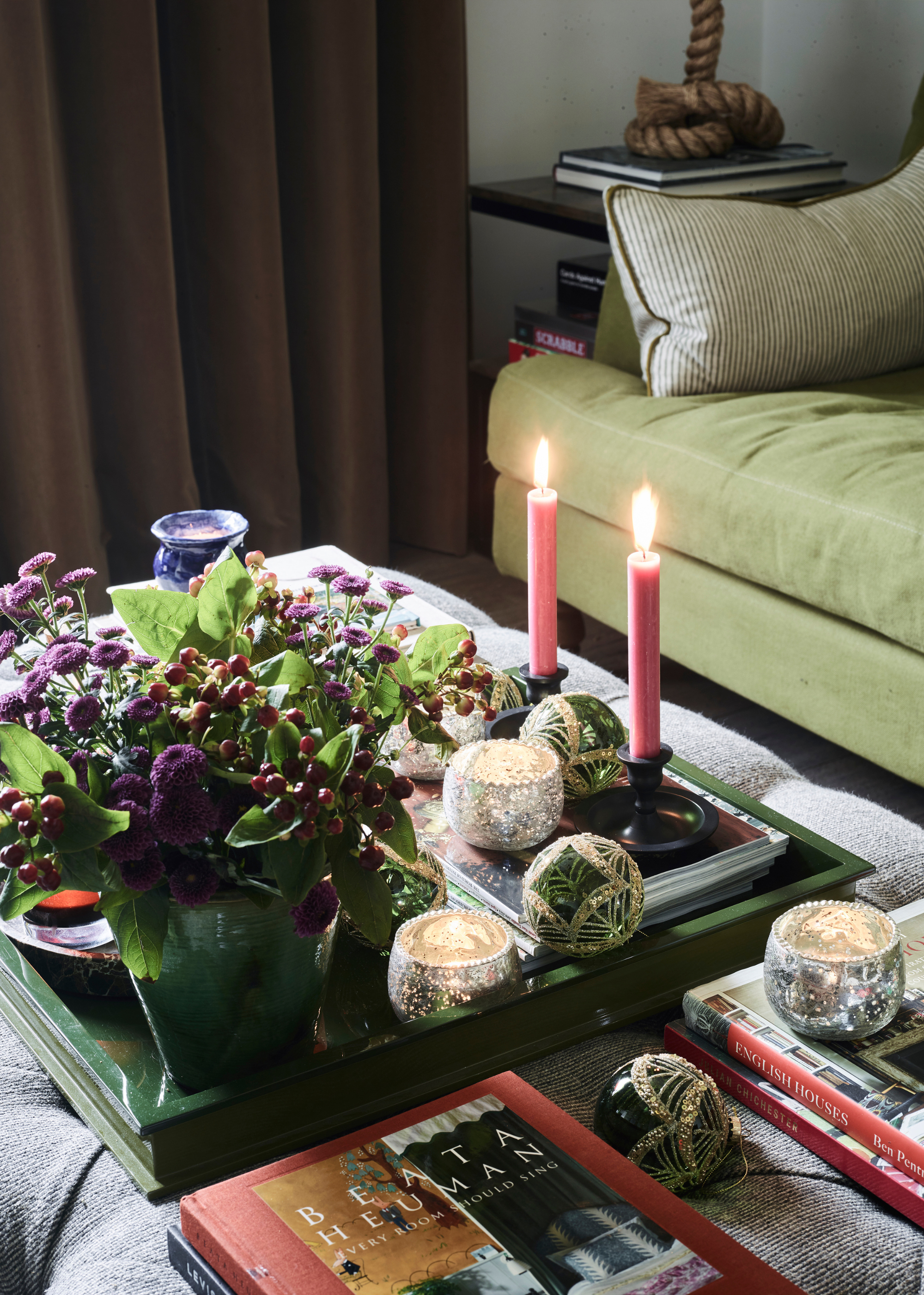 A button pouf coffee table is decorated with candles, baubles and winter berries and foliage in a vase on a green lacquered tree next to coffee table books