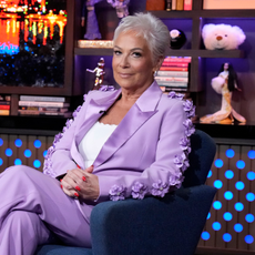 WATCH WHAT HAPPENS LIVE WITH ANDY COHEN -- Episode 22122. Denise Welch on set in lilac, floral suit, sitting on a blue armchair.