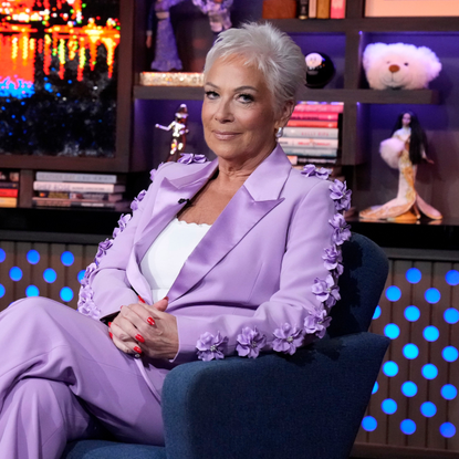 WATCH WHAT HAPPENS LIVE WITH ANDY COHEN -- Episode 22122. Denise Welch on set in lilac, floral suit, sitting on a blue armchair.