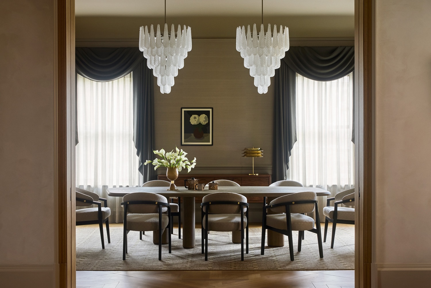 A symmetrical, sophisticated dining room viewed through a doorway. The space features a long wooden table surrounded by curved grey armchairs, two large tiered glass chandeliers, and tall windows with dramatic charcoal grey swags and sheer curtains