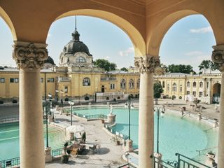 The Szechenyi Baths in Budapest built in 1913. Large outdoor blue water baths surrounded by historical buildings.