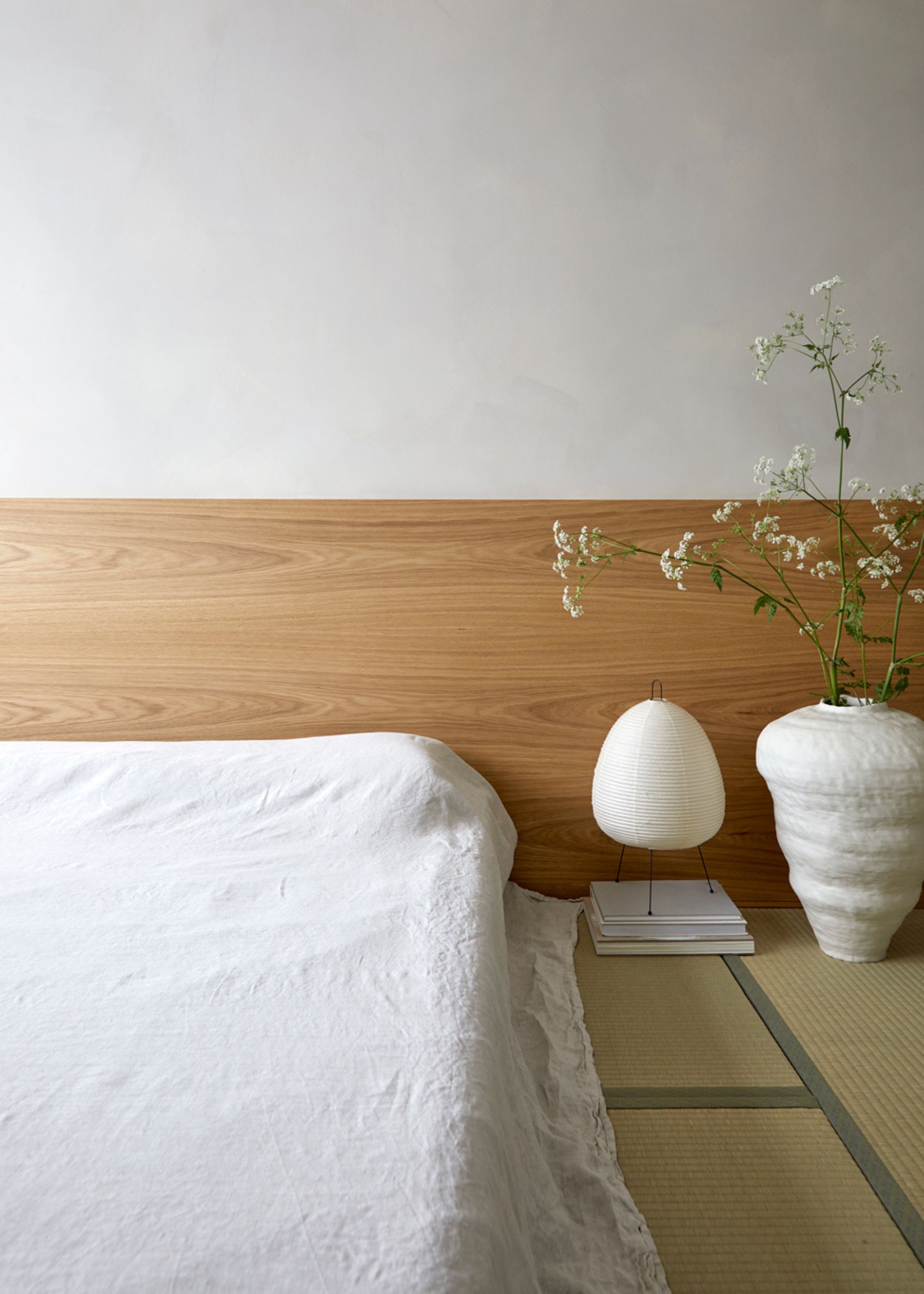 A traditional Japanese bedroom with tatami mats, a floor mattress, a paper lamp, a vase of flowers, and a wood headboard