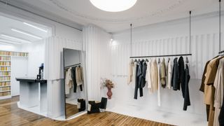 A modernly furnished fashion atelier features ceiling porthole windows, decor straddling rustic and industrial style in the tones of white and natural wood, and similarly hued garments on clothing rails hanging from the ceiling.