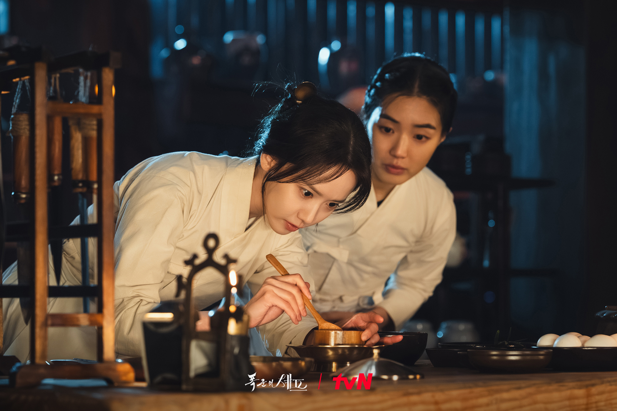 A woman (Lim Yoon-a) stirs a wooden spoon in a small bowl as another woman (Yeon Seo-ah) peeks over her shoulder, in &#039;Bon Appetit, Your Majesty.&#039;