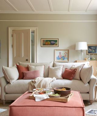 A white living room with a white couch, coral pink ottoman, and pink throw pillows