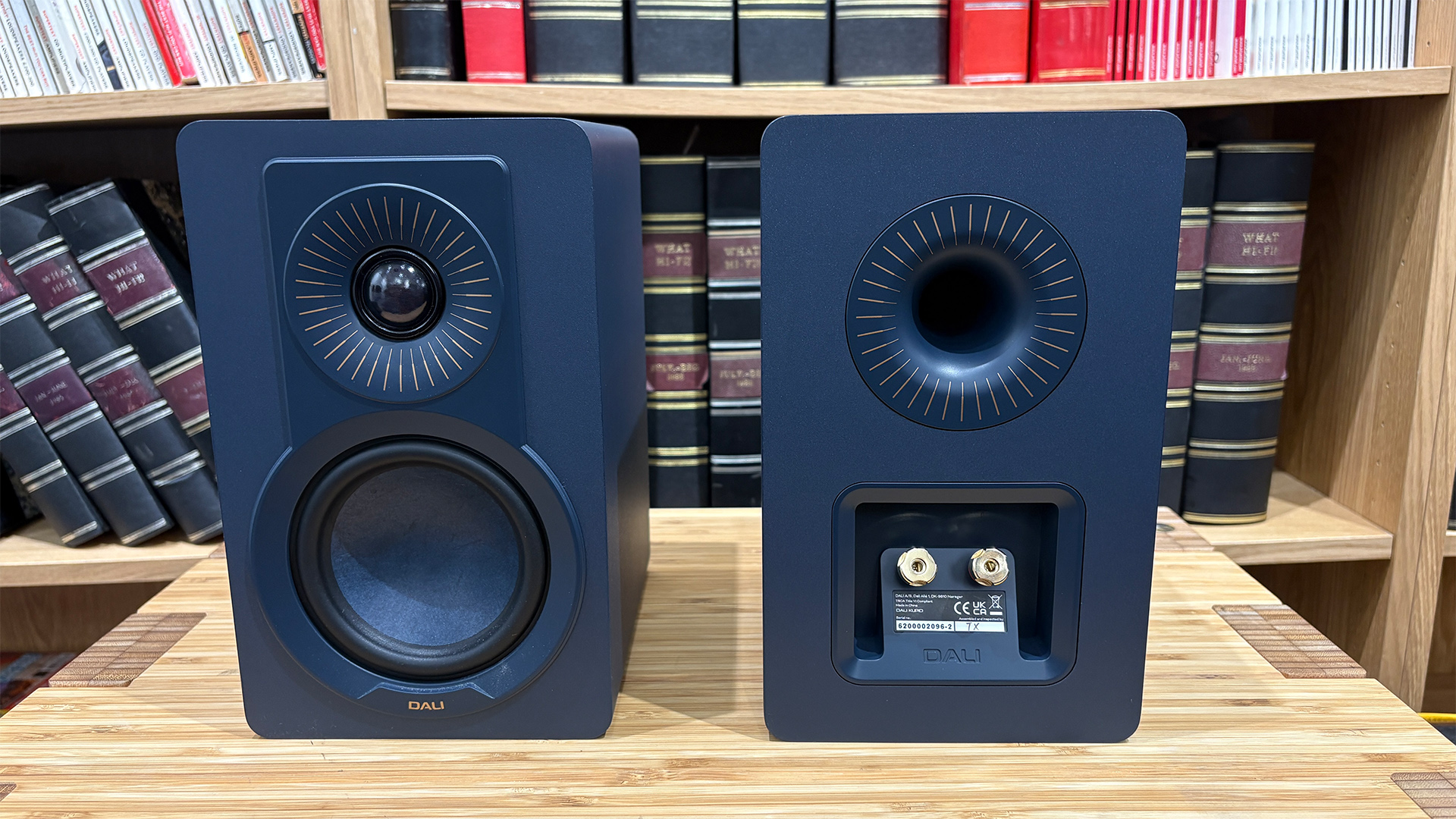 Dali Kupid standmount speakers on wooden hi-fi rack in front of bookcase, one is turned around to show binding posts