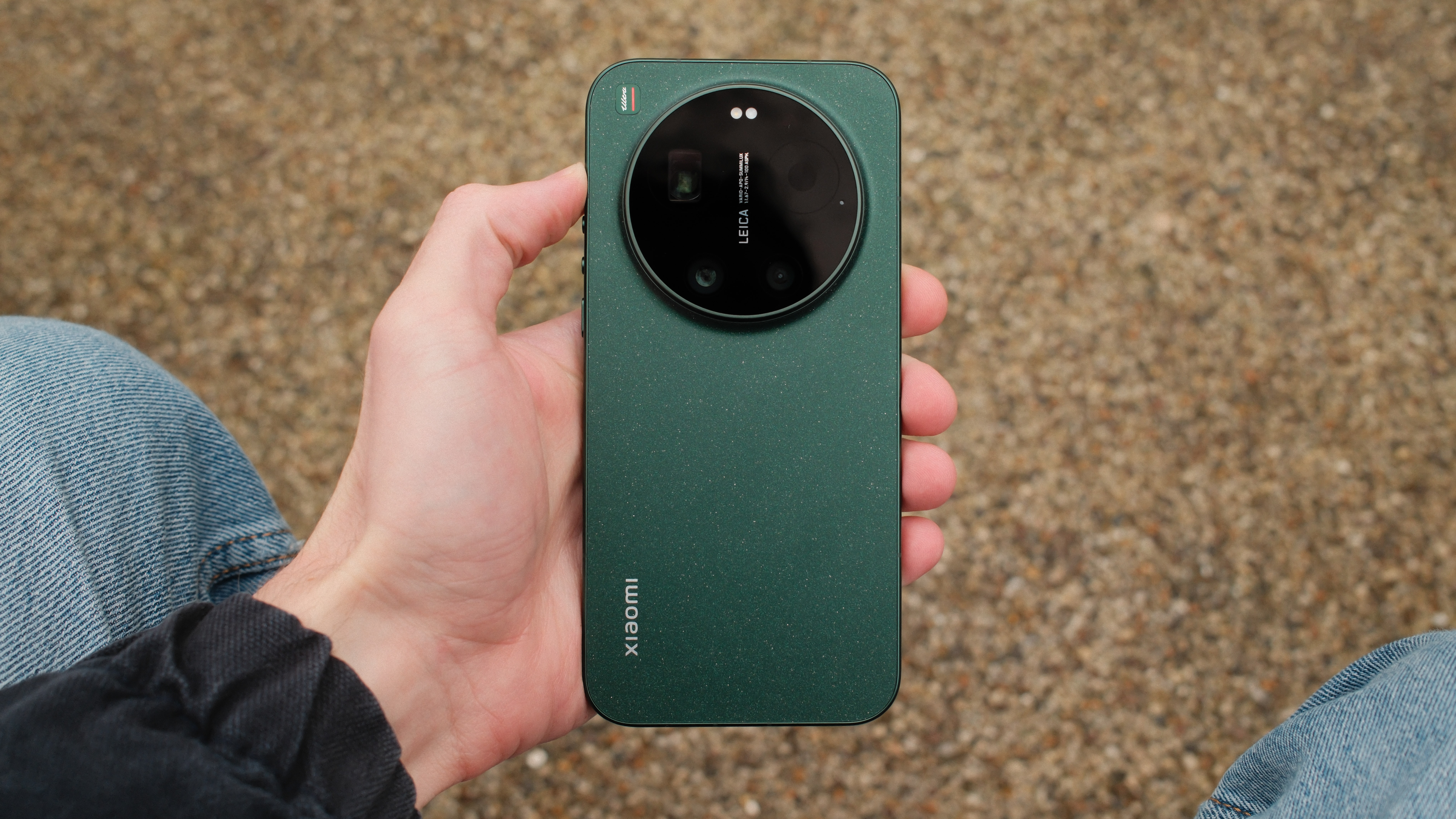 Rear view of the green Xiaomi 17 Ultra smartphone showing its oversized circular Leica-branded camera bump, held above a gravel surface.