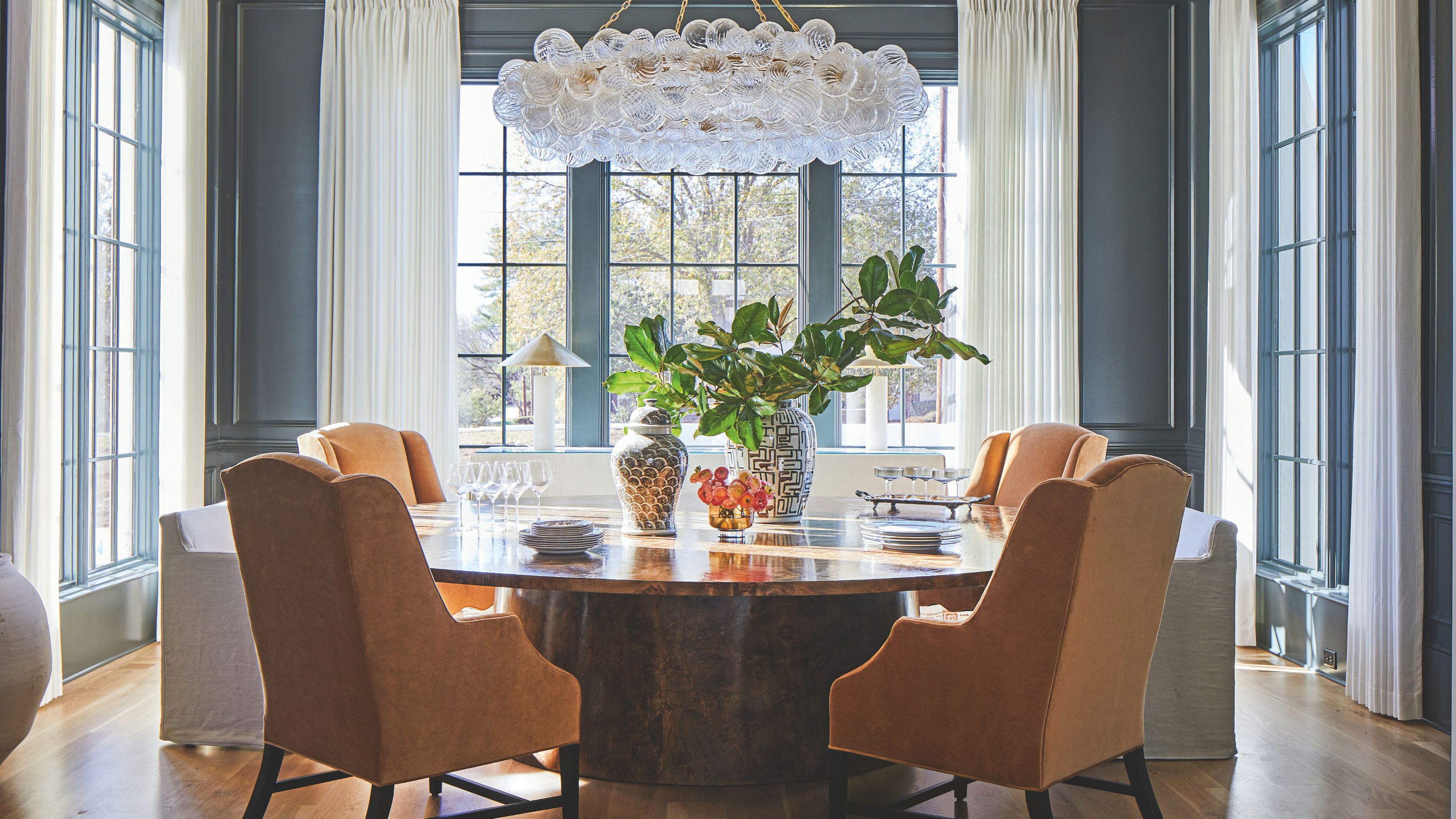 A dark blue/gray dining room with large windows and long white drapes. A large wooden dining table sits in the middle with a chandelier above and orange upholstered dining chairs around