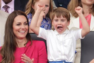 Princess Kate wearing a bright pink dress sitting down and laughing at Prince Louis who is wearing a white shirt and blue shorts, standing up and cheering with his tongue out