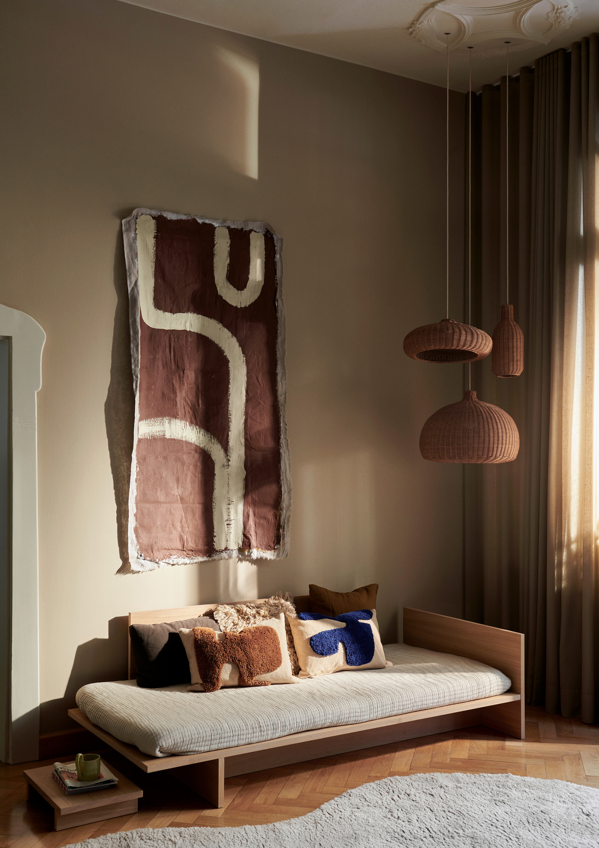 A cozy living room with a muted color scheme; there is a fabric wall art on the warm beige walls above a low daybed with scattered throw pillows with blue and brown accents, a curved gray rug on a wooden herringbone floor and three woven pendant ceiling lights in a warm brown in different shapes and at different heights