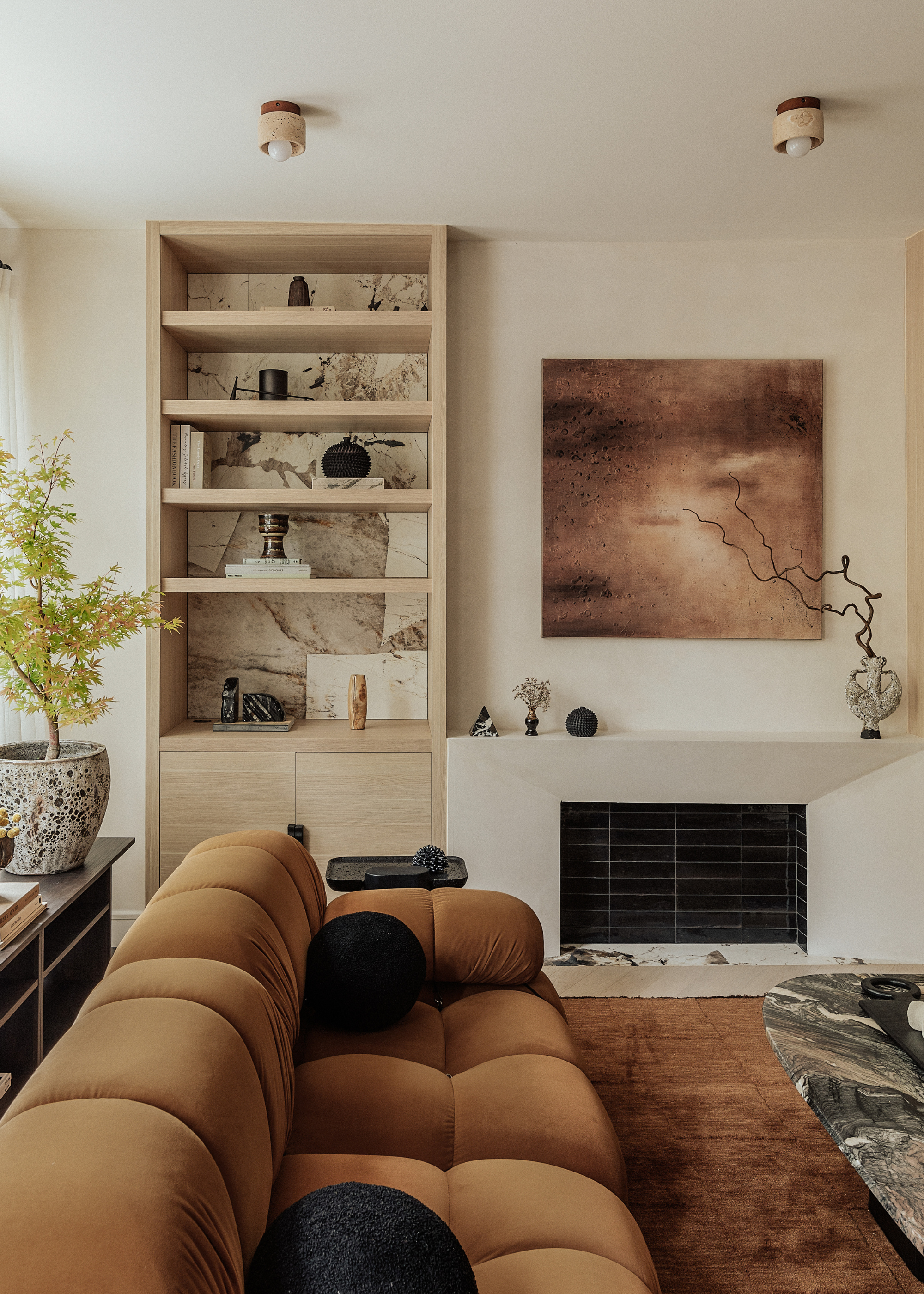 A warm white living room with a cinnamon couch, a plush area rug, a stone coffee table, a potted tree in a textured planter on a sideboard, a walled fireplace, art on the wall and a wood unit with books, vases and bowls.