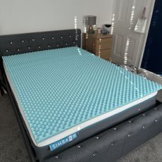 The turquoise blue Aidapt Memory Foam Mattress Topper on top of a mattress on a grey upholstered bed in a bedroom. The surface of the mattress topper has an egg-crate texture