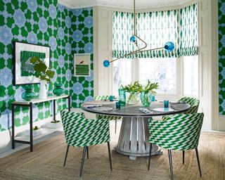 Colorful dining room decorated in blue and green, blue and green floral wallpaper, dark wood rounded dining table, four upholstered dining chairs in blue and green zig-zag pattern, natural texture rug, metal and blue glass sculptural hanging pendant