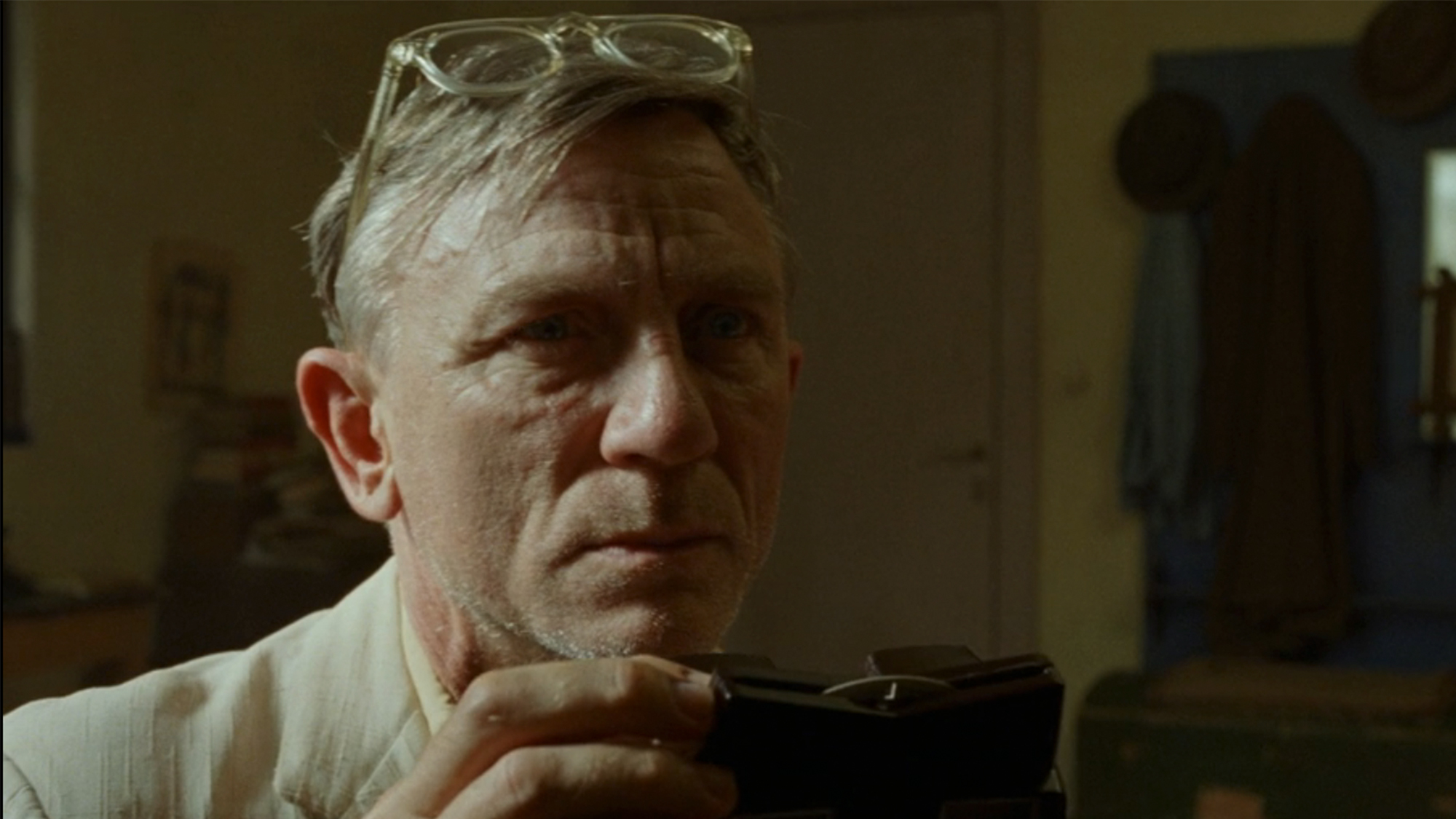 A close up of Daniel Craig with glasses on top of his head, holding a camera in Queer