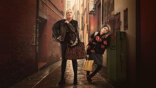 Daisy May Cooper as Costello and Fleur Tashjian as Iris stand in an alley holding bags in Rain Dogs
