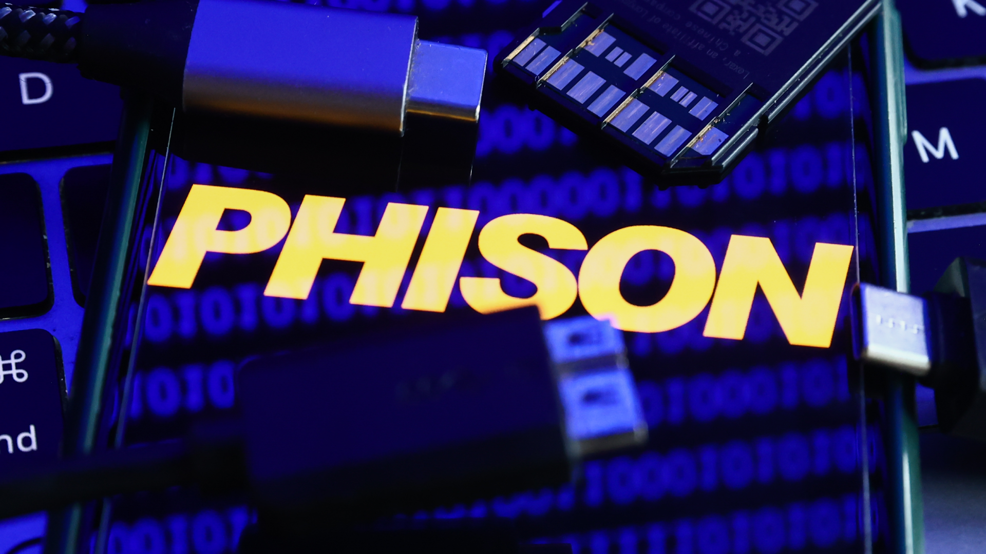 Phison logo displayed on a phone screen with a binary code reflected on it, a laptop keyboard, a memory card, an adaper and cables are seen in this illustration photo taken in Krakow, Poland on January 30, 2023.